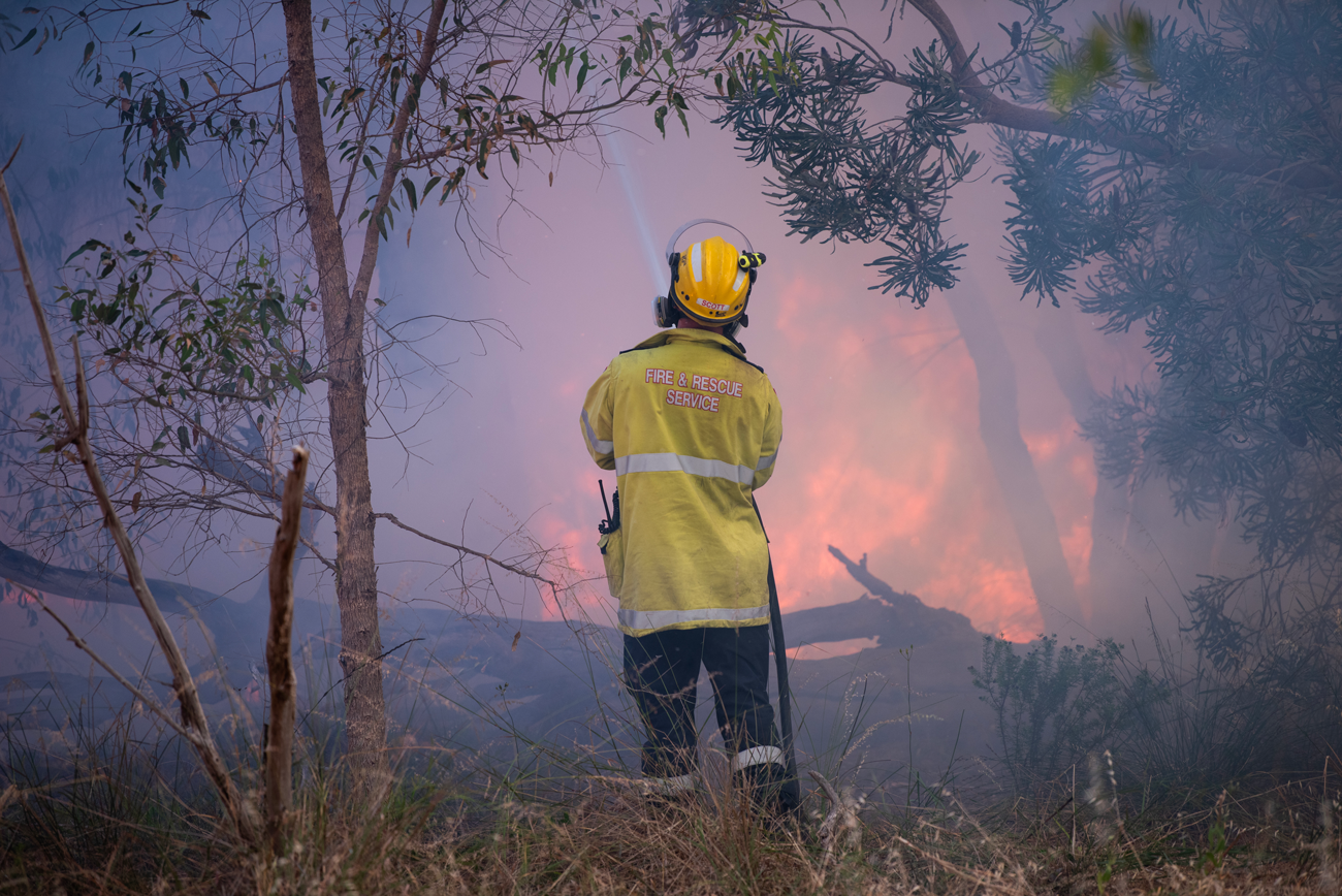 A firefighter stands in the middle of the frame with a hose with a burning fire in the background.