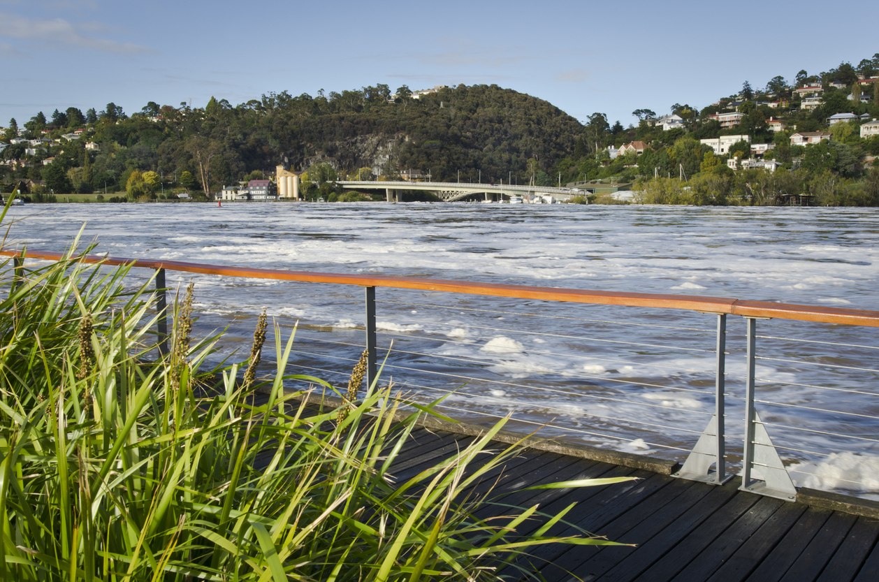 A walkway and fence with rushing water and a bridge and mountains behind it.