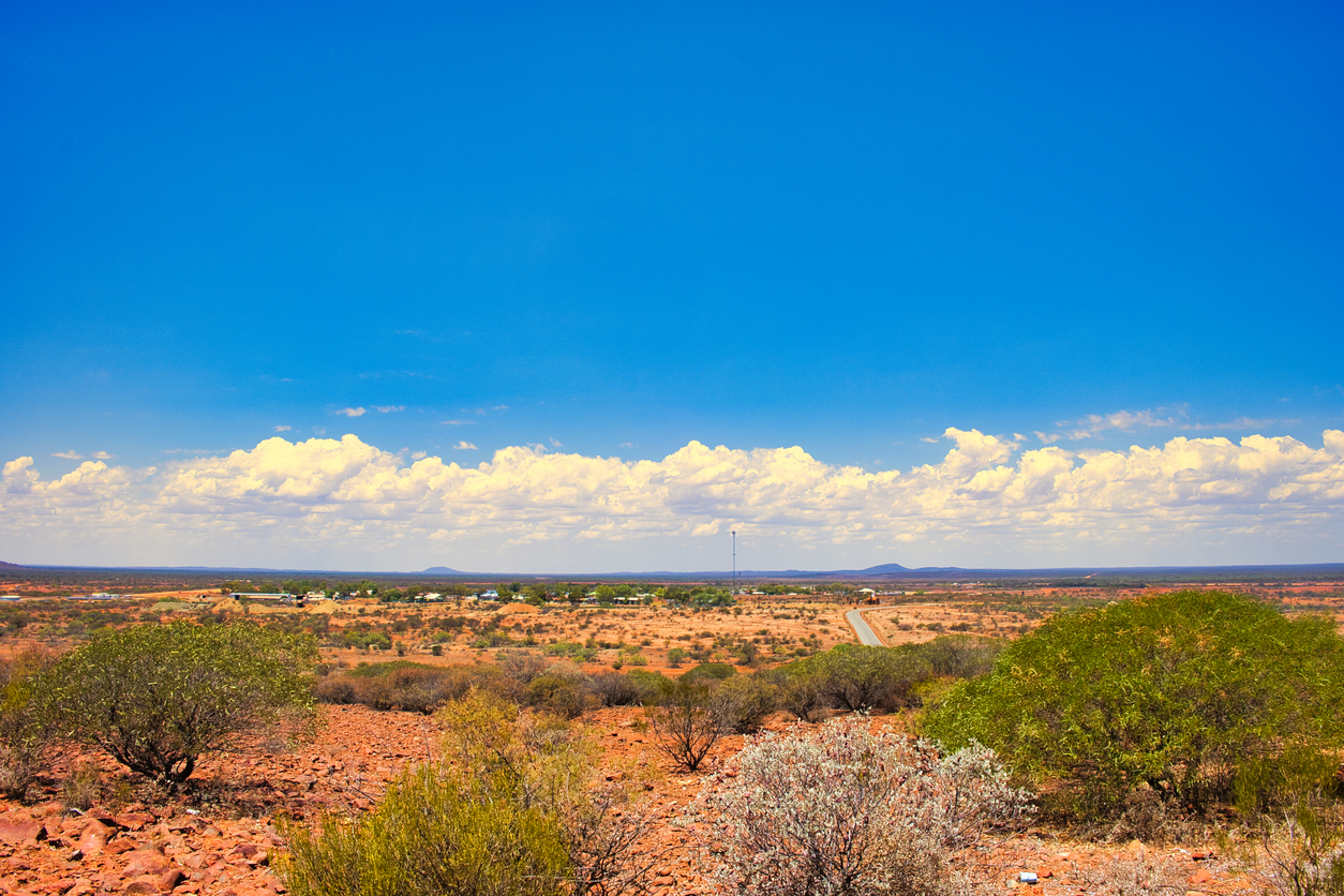 A dry red landscape with some green shrubs and a blue sky.