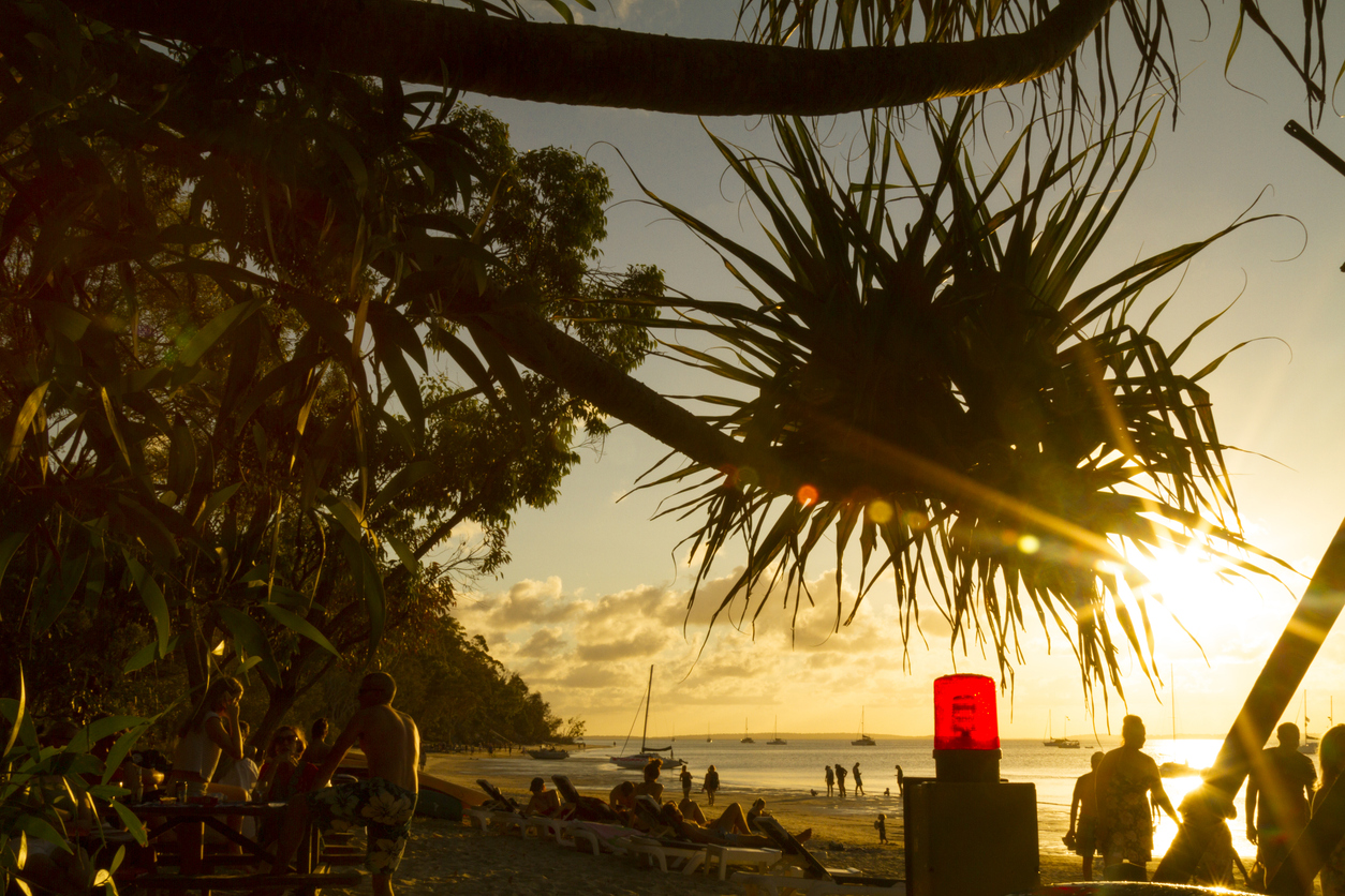 A busy beach with pandanus tree shadow caused by setting sun. A red warning beacon is visible.