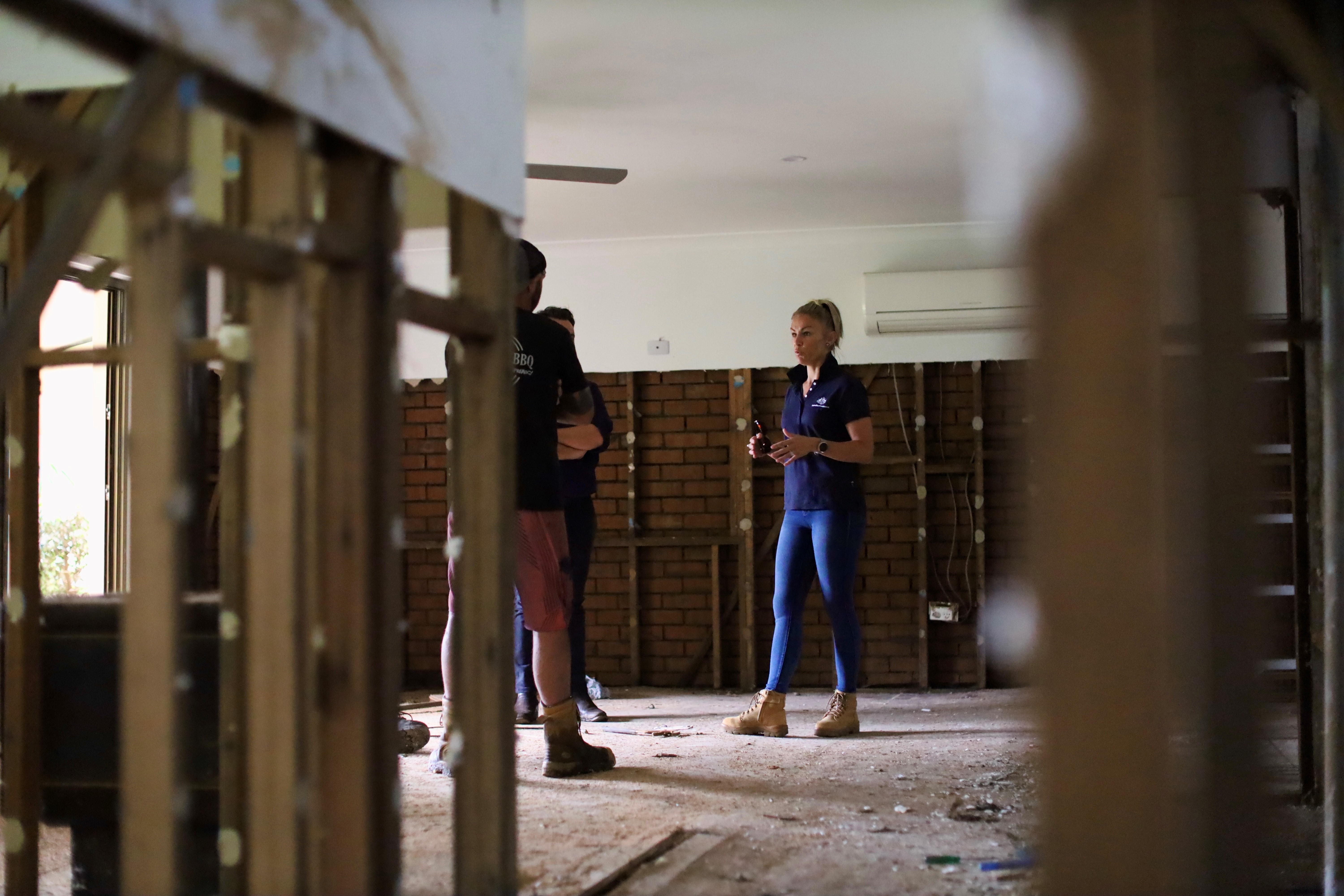 A NEMA employee in a blue outfit and work boots talks to two people in a disaster impacted room with exposed wooden beams and brick walls.