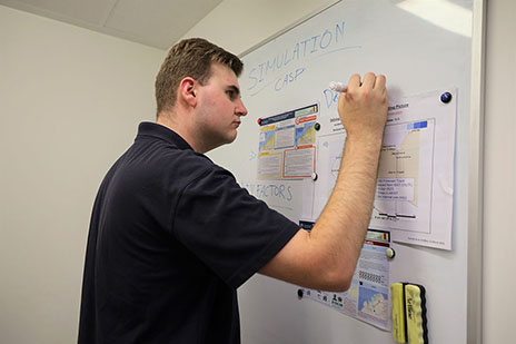 A person in a navy polo shirt with NEMA branding standing on the left of a whiteboard. They’re holding a whiteboard marker.