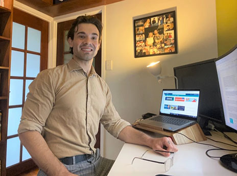 A person in a beige shirt is sitting in a home office in front of a laptop and smiling.