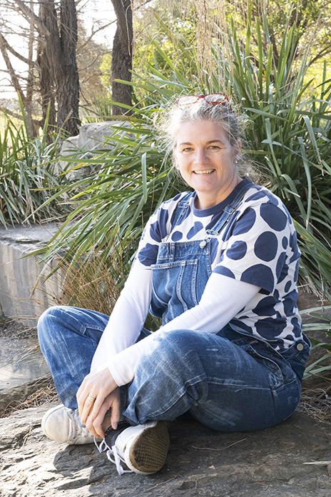 A person in a white top with large navy dots and denim overalls is sitting on the ground in front of green plants and smiling.