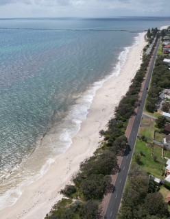 An aerial view of a coastal land with a sandy beach lined on the left side, with residential neighbourhoods on the right. The shoreline is bordered by gentle waves, with the ocean extending to the horizon. 