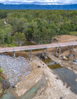 A newly constructed bridge goes over a river. Building equipment is under the bridge as workers in high vis move around the worksite.  