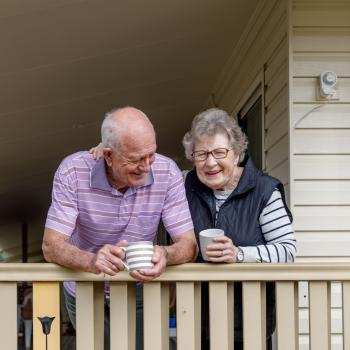 An elderly couple drink tea on their home veranda while smiling.