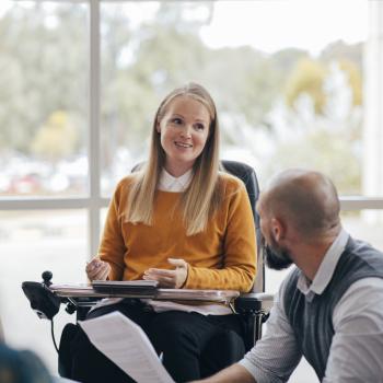 A woman looks happy and confident as she leads a group discussion at her place of work. She is a wheelchair user and has Muscular Dystrophy.