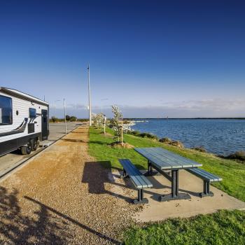 A caravan parked by a park bench, some grass and a large body of water.