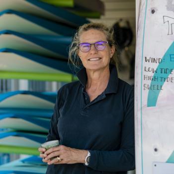 A woman stands in the doorway of a surf shop. Surfboards are stacked to the left.