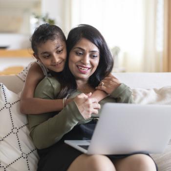 A woman sitting on the couch using a laptop is comforted by her child giving her a hug around the neck.