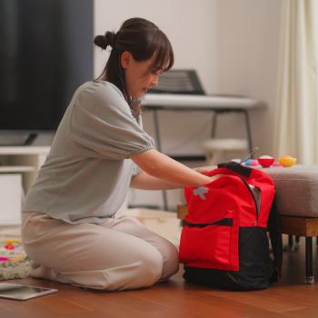 A woman kneels on the ground in a living room placing items into a first aid backpack.