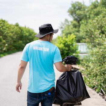 A person wearing a volunteer shirt walks down a road with a trash bag.