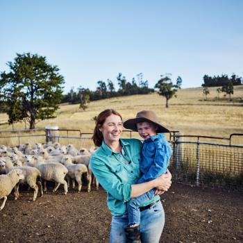 A farmer and a child embrace on a property with sheep in the background.