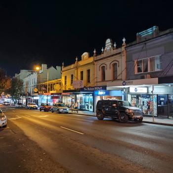 A street at night with shops and cars parked by the side of the street.