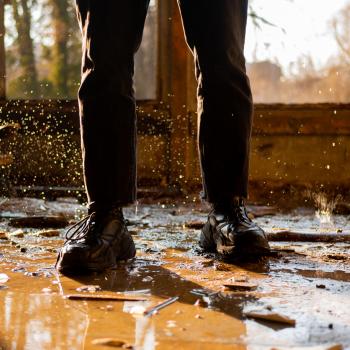 Boots stand inside a house with a muddy floor and debris.