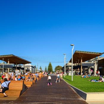 People sit an a refurbished community area with green grass and large wooden seating.