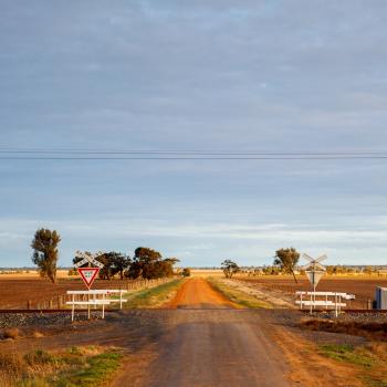 A rail crossing in a rural area with a red dirt road on the other side.