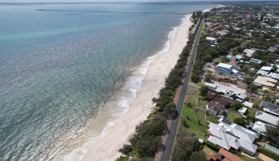 An aerial view of a coastal land with a sandy beach lined on the left side, with residential neighbourhoods on the right. The shoreline is bordered by gentle waves, with the ocean extending to the horizon. 