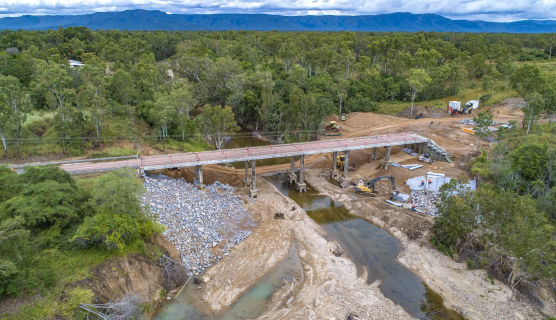 A newly constructed bridge goes over a river. Building equipment is under the bridge as workers in high vis move around the worksite.  