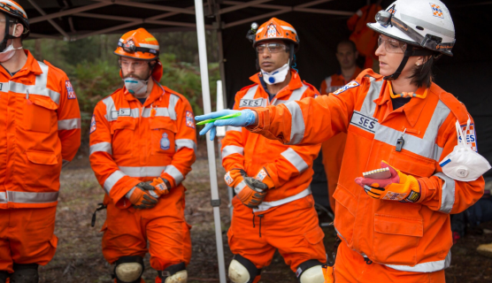 SES volunteers stand engaged in conversation. A woman stands at the front of the crowd pointing to something out of frame. She is wearing safety equipment (glasses, a white helmet and and gloves). The men in the background also wear safety equipment including white knee pads and masks. 