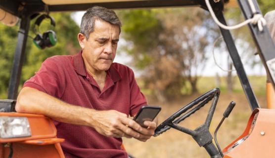 An elderly man sits on a tractor looking at his mobile phone while wearing a red top. Brown grass is out of focus in the background. 