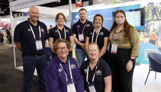 Six people from the National Emergency Management Agency (NEMA) pose for a photo in front of a NEMA-branded booth at a conference. They are wearing event lanyards and uniforms.