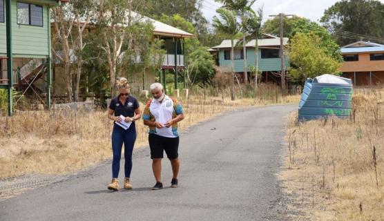 Two people walk down a street. Damaged houses are visible in the background.