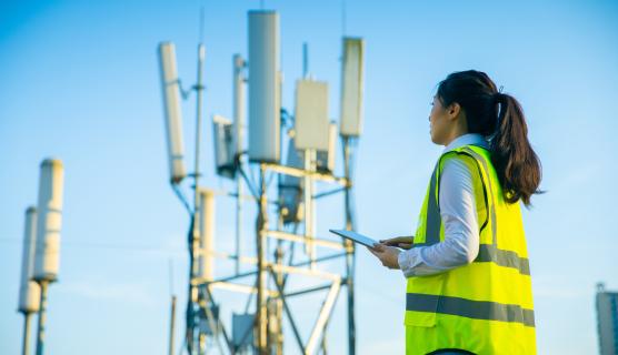 A woman wearing a high-visibility vest is holding an iPad. In the background is a radio signal tower set against a clear blue sky.