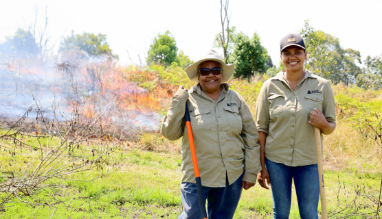 Two people in khaki uniforms and denim jeans stand in front of a controlled burn.