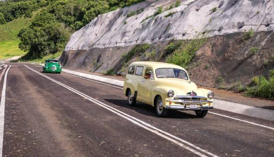 Two historical cars drive down a recently repaved road with large wall to the right.