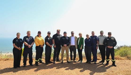 A group of emergency management experts standing together outdoors on a hilltop overlooking the ocean, wearing various clothing and footwear, with a clear sky in the background.