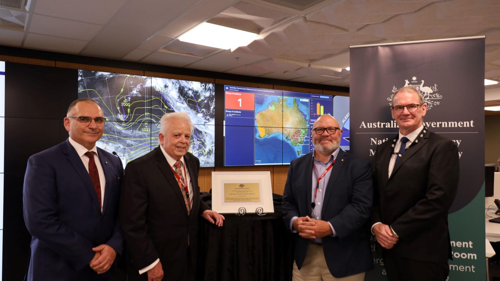 Brendan Moon, Joe Buffone, Rob Cameron and David Templeman posing in front of the digital screen of the National Situation Room.
