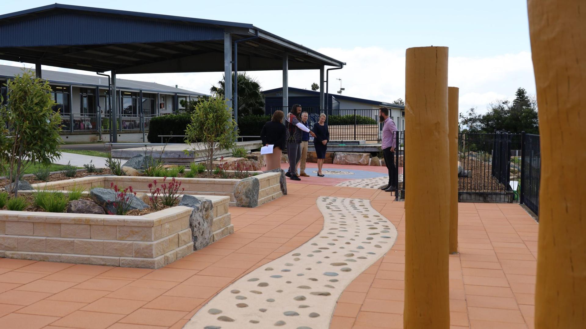 Paved open area with two wooden posts in foreground with garden beds protected by an aluminium awning on left hand side, with a concrete path in centre of image.