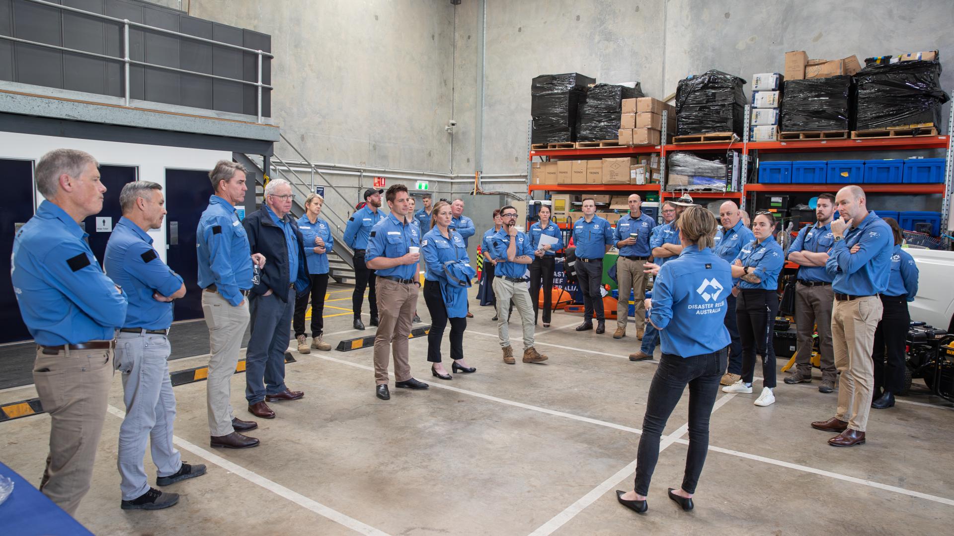 A crowd of people in Disaster Relief Australia uniforms stand in a new warehouse space listening to a speaker. 