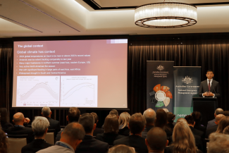 A speaker stands at a lectern on a stage with NEMA pull up banners beside them. A large screen shows text about the global climate context. 