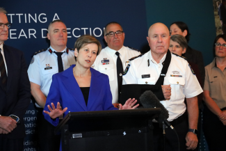 A woman stands behind a lectern speaking while gesturing with her hands.