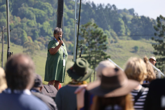 A woman stands on stage outside speaking into a microphone in front of a crowd.