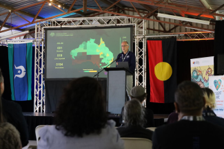 A person stands at a lectern speaking to an audience. Beside them is a large Australian Aboriginal flag.