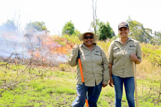 Two people in khaki uniforms and denim jeans stand in front of a controlled burn.