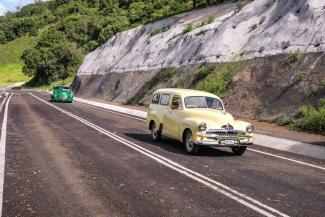 Two historical cars drive down a recently repaved road with large wall to the right.