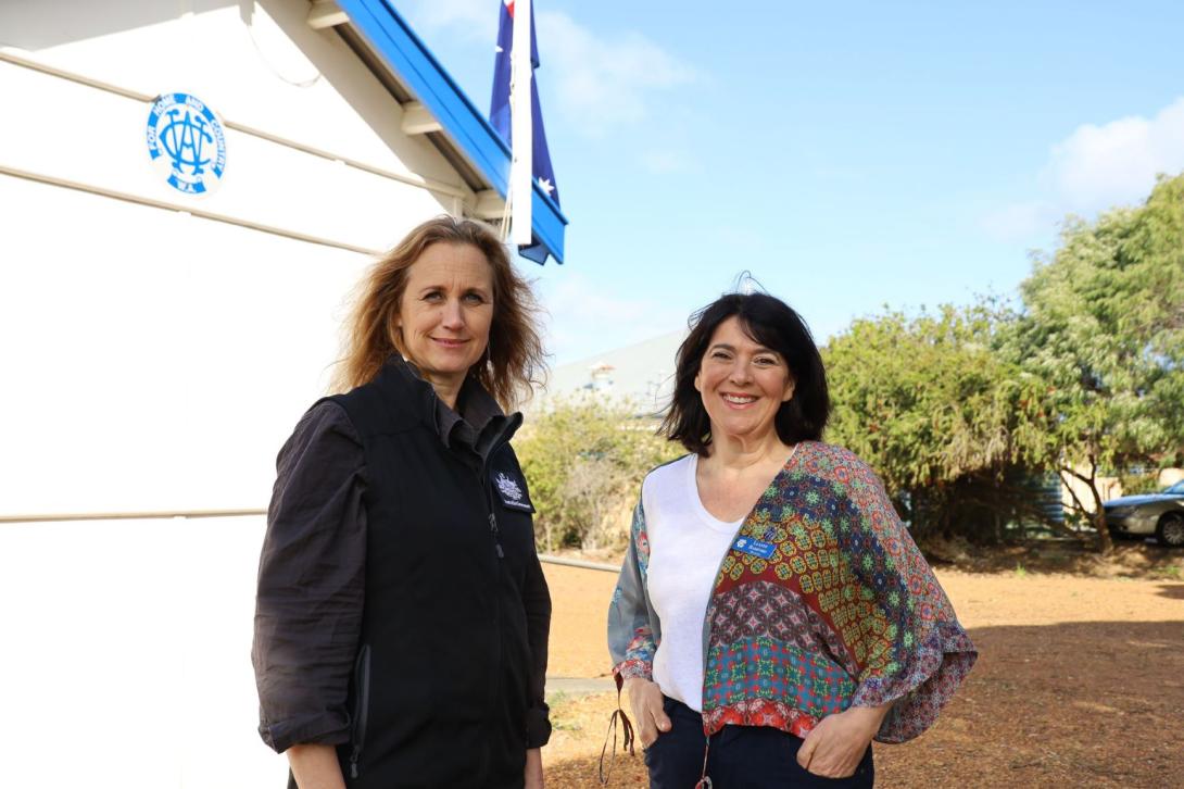 Two people facing camera taken outside next to a white wall exterior of a building. One person on the left wears a branded shirt featuring the crest of the Australian Government and National Emergency Management Agency branding the other person on the right is wearing a white and multicoloured top.
