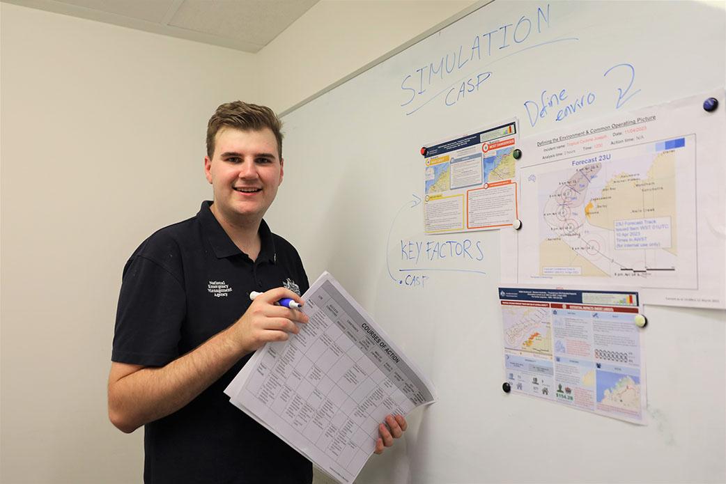 A person in a navy polo shirt with NEMA branding standing on the left of a whiteboard. They’re holding a whiteboard marker and a paper folder and are smiling. 