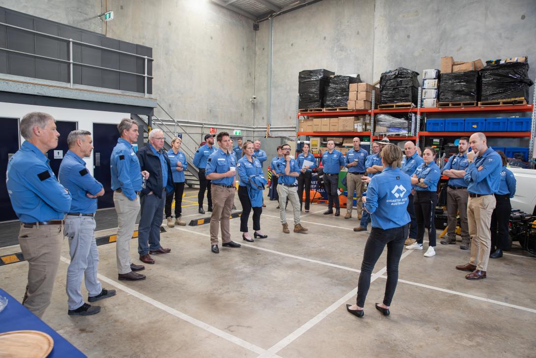 A crowd of people in Disaster Relief Australia uniforms stand in a new warehouse space listening to a speaker. 